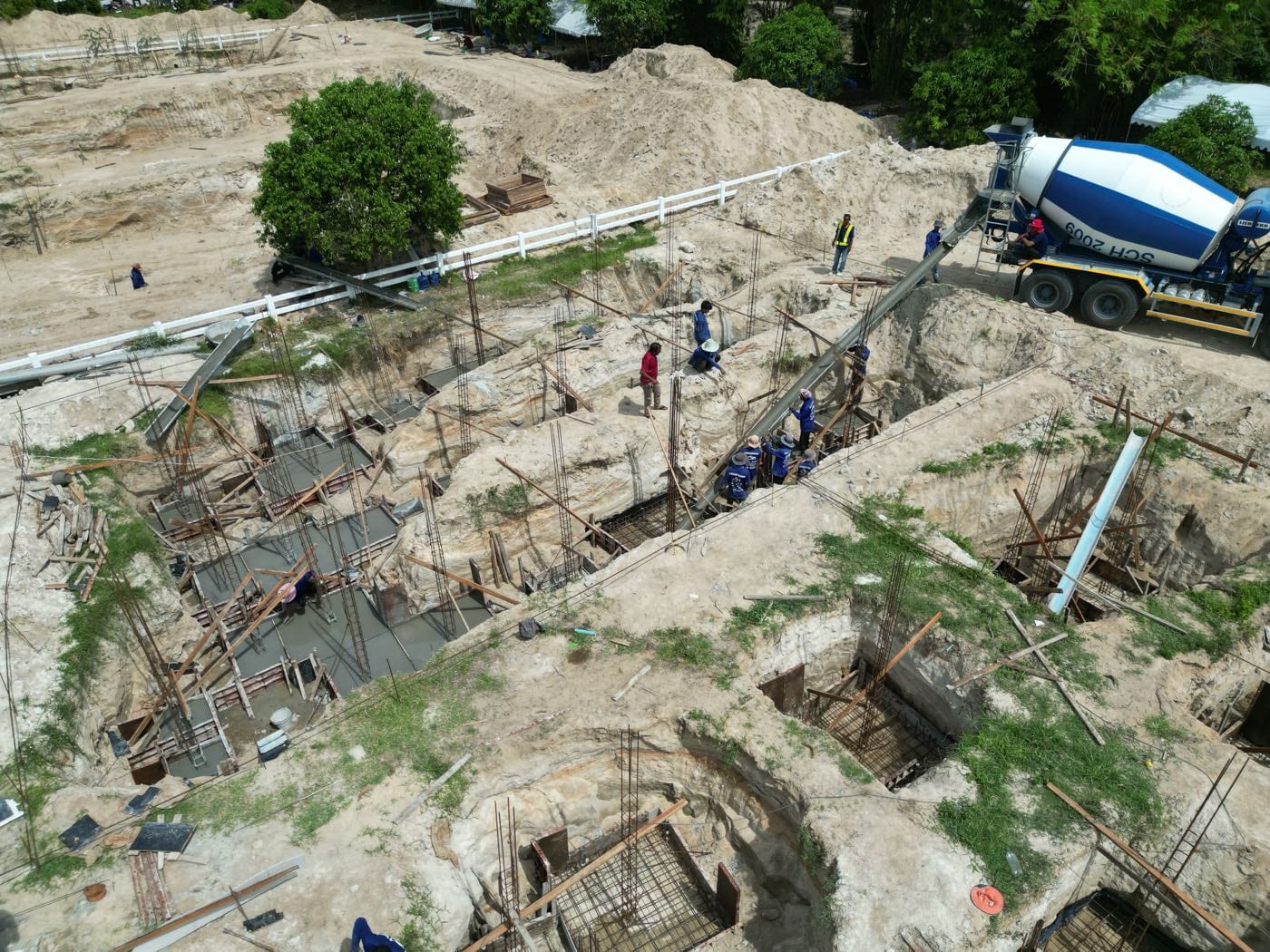 Foundations under way: excavated trenches with steel reinforcement cages, a concrete mixer truck with its chute extended and a crew placing concrete