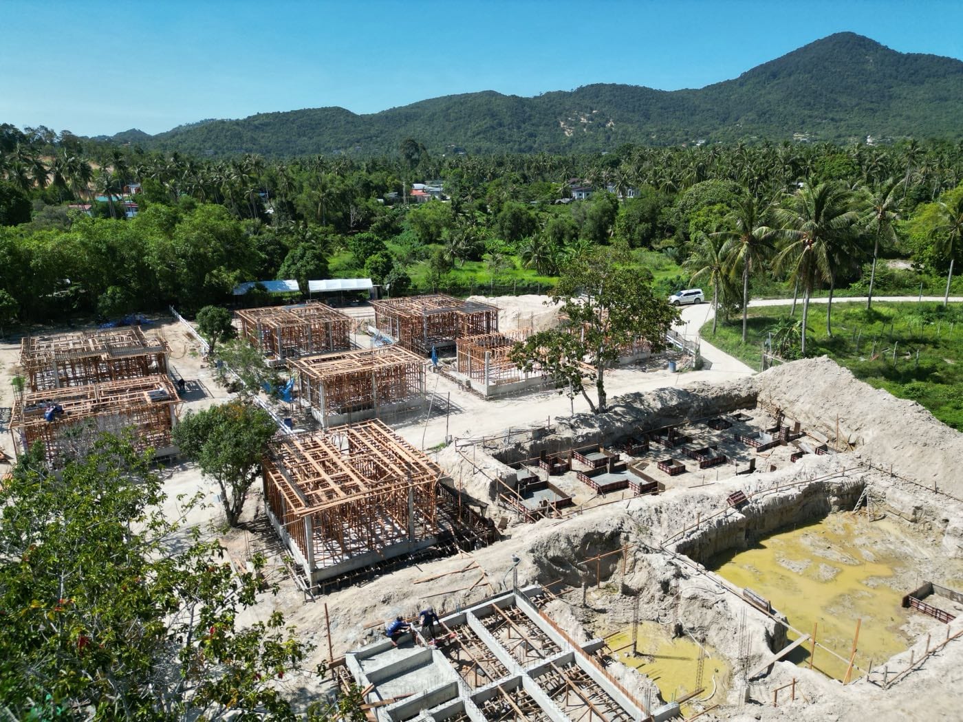 Houses in timber formwork with structural frames going up across the site, the wellness hub excavated alongside and the Koh Phangan ridge line behind